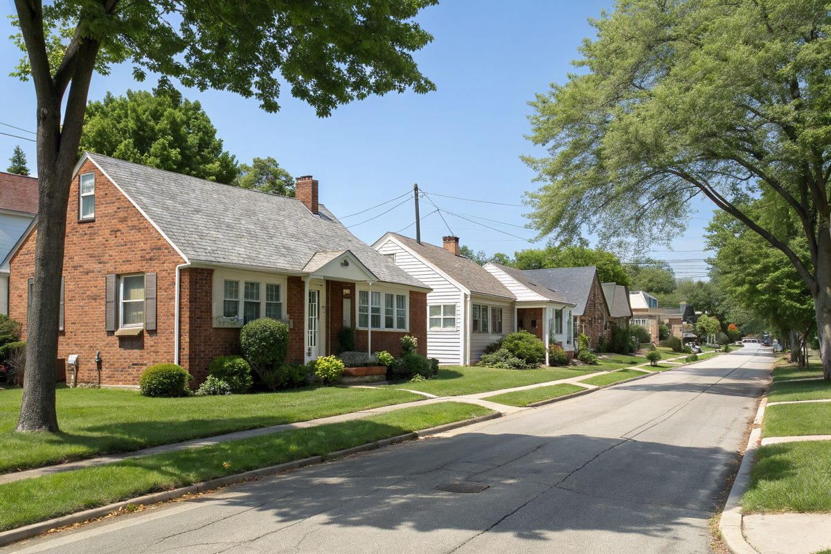 A street of small brick and frame Cape Cod houses with gray shingle roofs under big shade trees